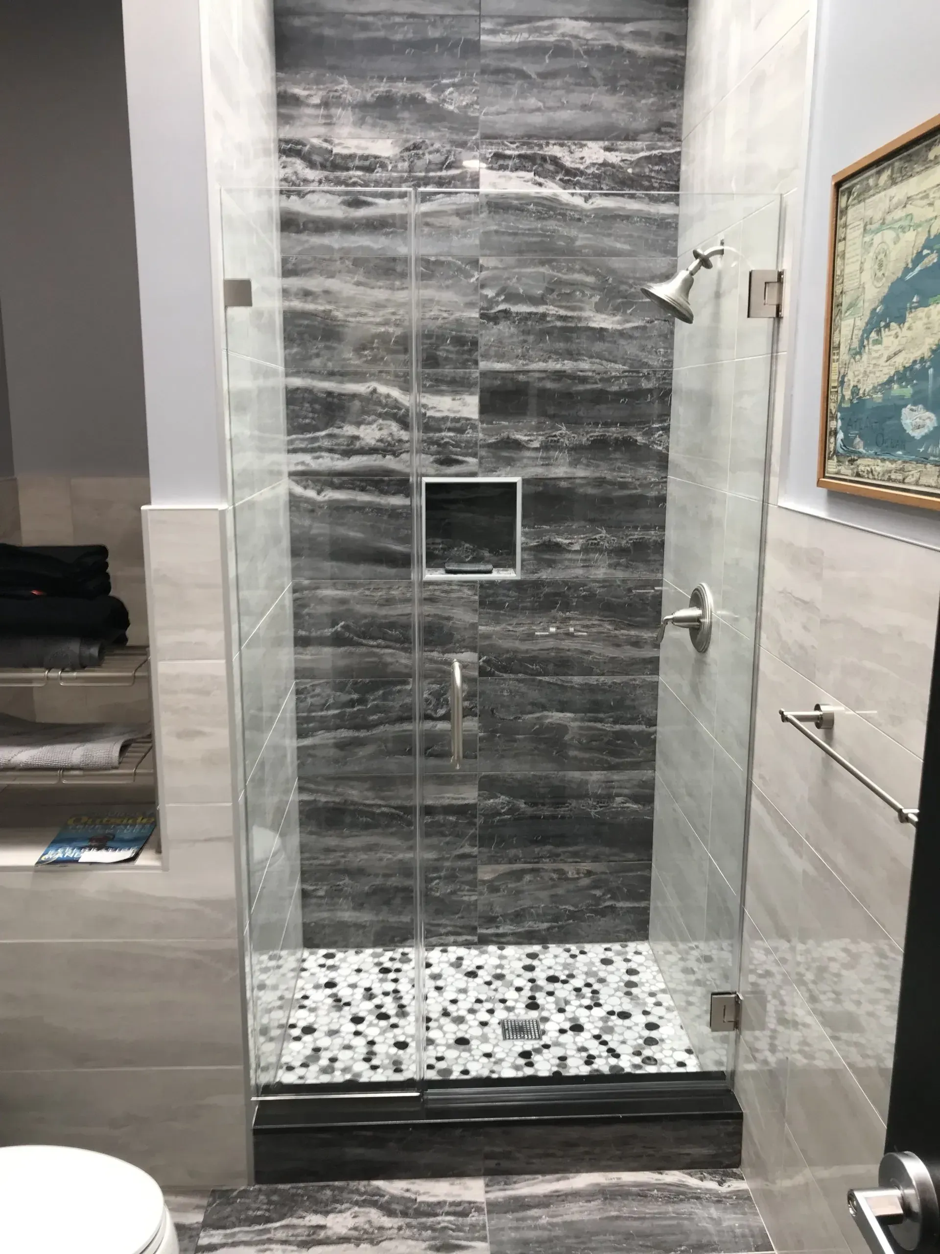 Shower with gray and white marbled tile, glass door, pebble floor, and silver fixtures.