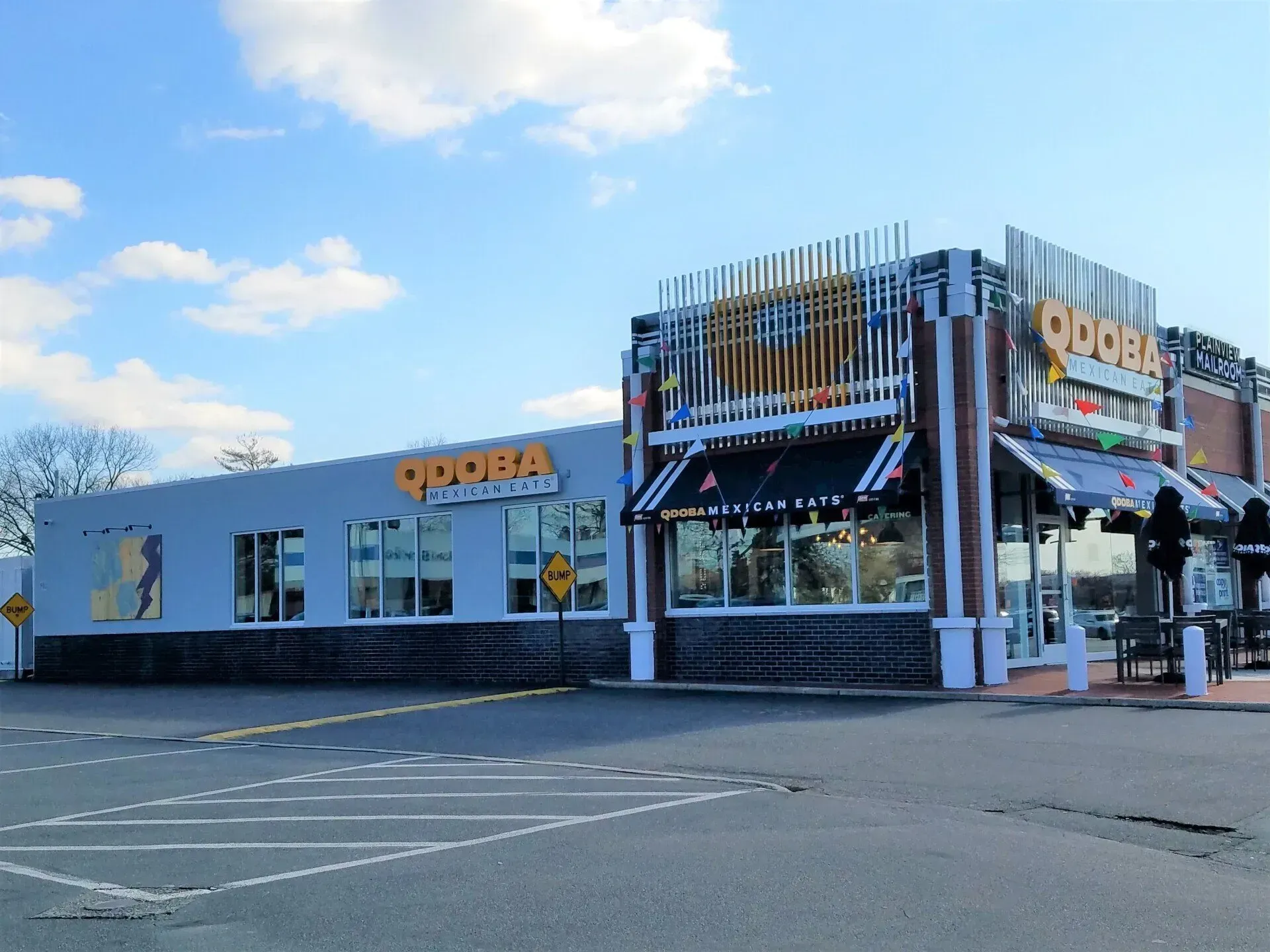 Exterior view of a Qdoba restaurant on a sunny day. Gray and brick building with awnings and yellow signage.