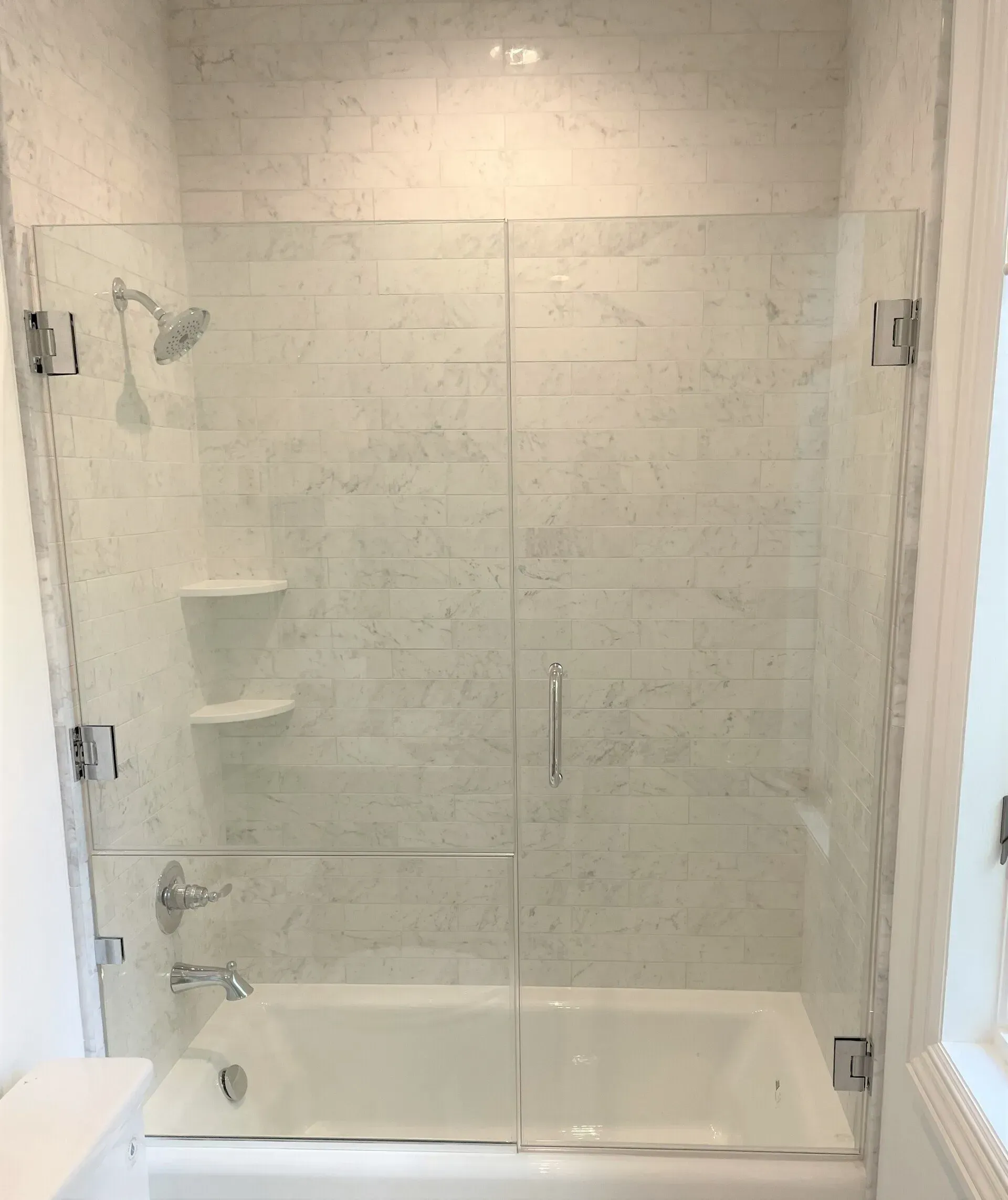 Glass shower enclosure over a white bathtub with marble tile walls and chrome fixtures.