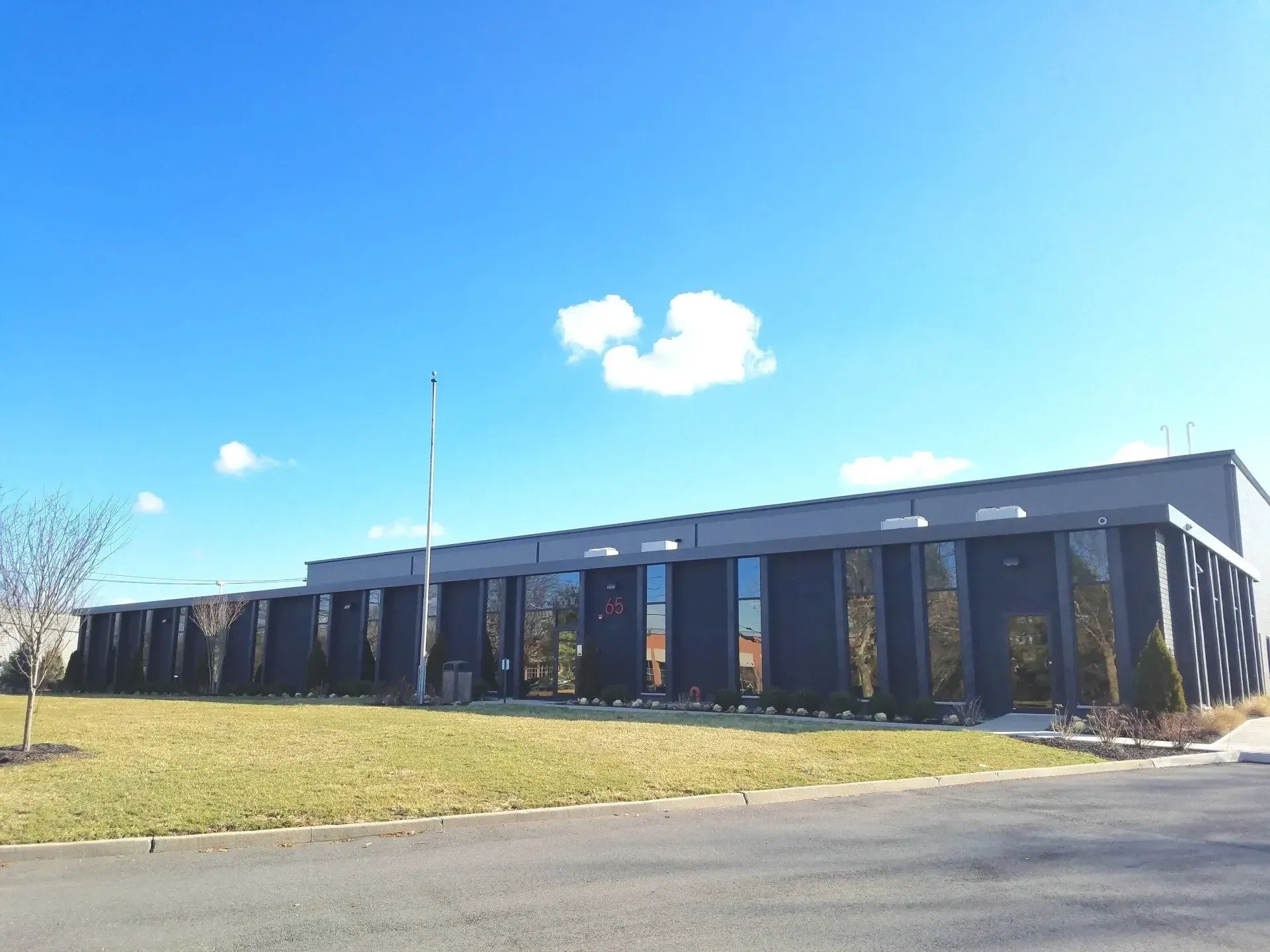 Modern, dark-colored building with large windows, set against a blue sky with a few clouds; grass and bare trees in foreground.