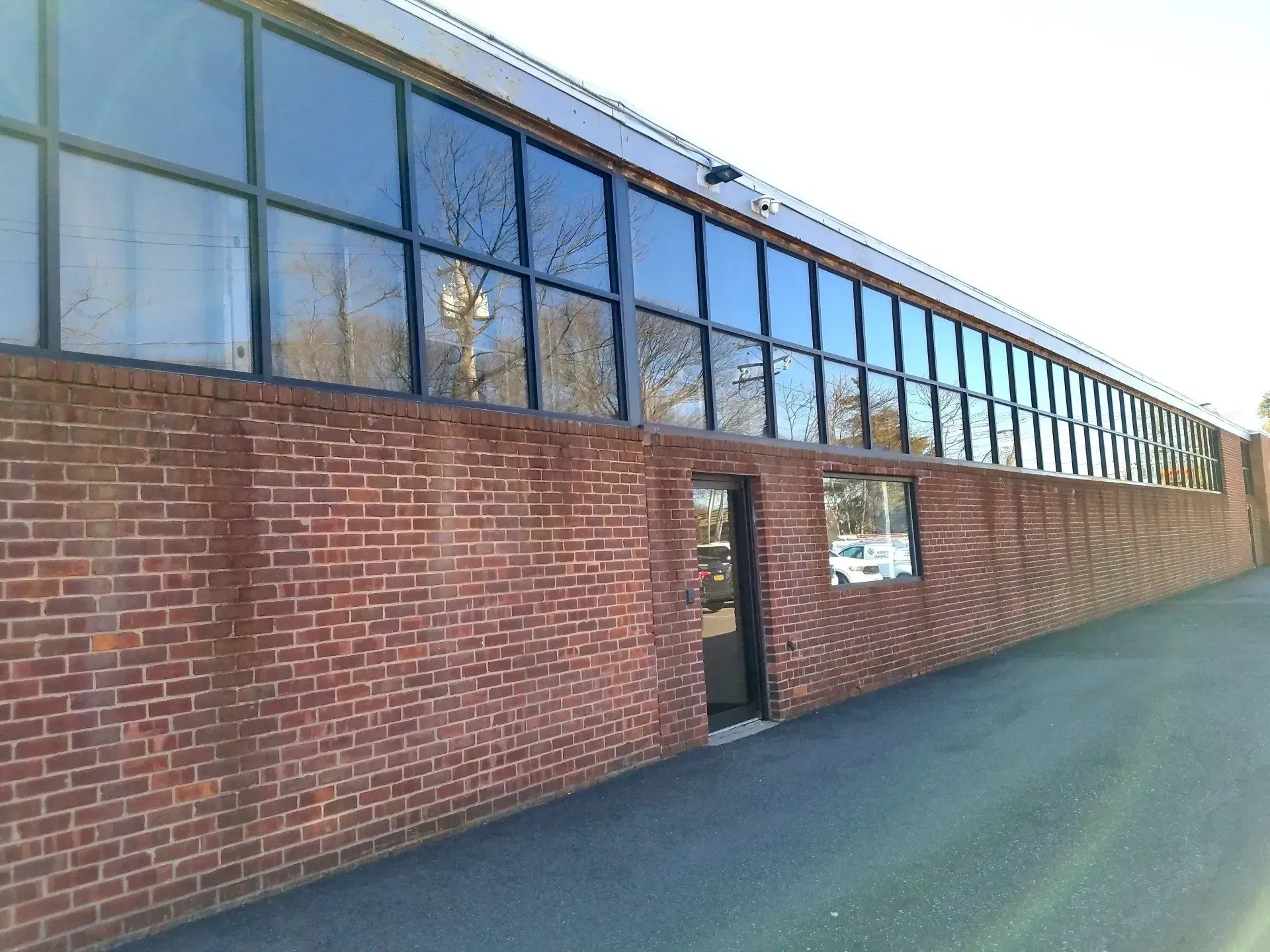 Red brick building with long row of windows above, dark door, asphalt pathway.