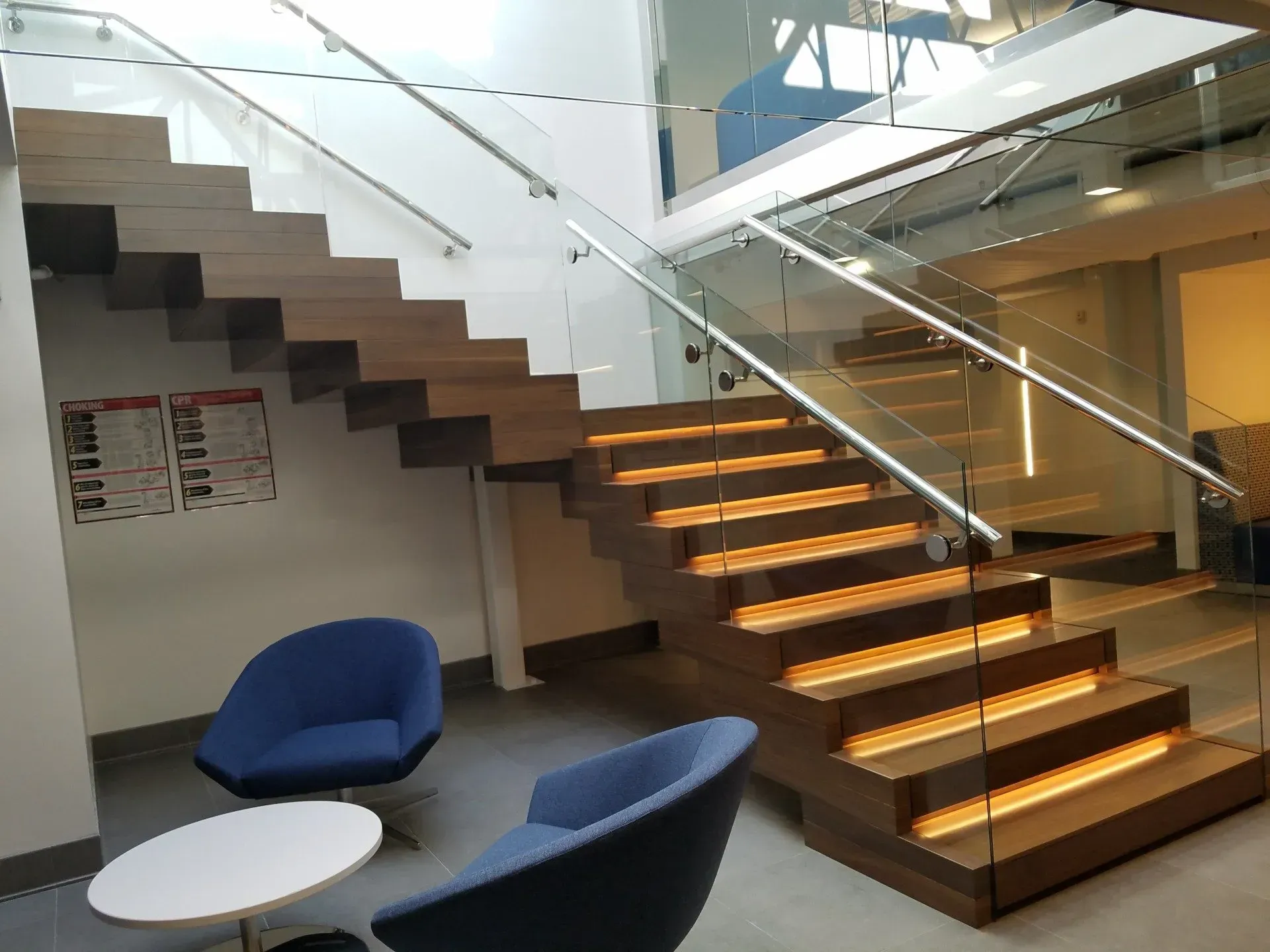 Wooden staircase with glass railing and built-in lighting, leading to upper level. Two blue chairs and white table in foreground.