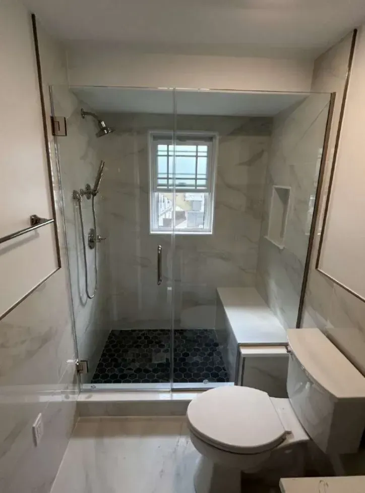 Modern bathroom with glass shower, marble tile, bench seat, and toilet.