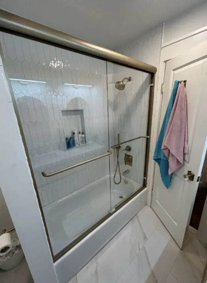 Bathroom with glass shower door, white tiled walls and floor, and towels hanging on the door.