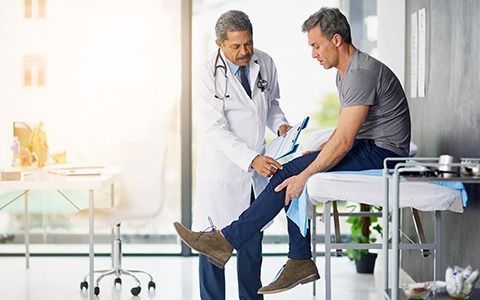 Doctor examining a patient's knee in a bright office. The doctor holds a chart.