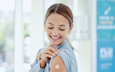Woman smiles, touches bandage on her arm after receiving a vaccine; indoors.