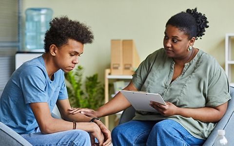 Counselor comforts teen. Woman in green shirt, man in blue shirt. Both seated, indoor setting.