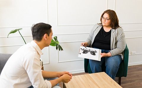 Man in therapy with a therapist holding a Rorschach inkblot test in a neutral-colored room.