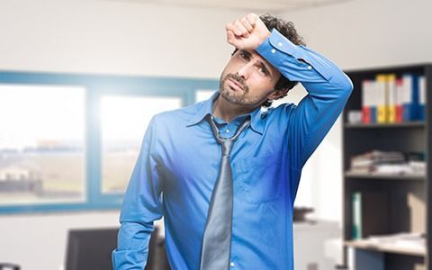 Man in blue shirt sweating, wiping brow. Appears uncomfortable in an office.