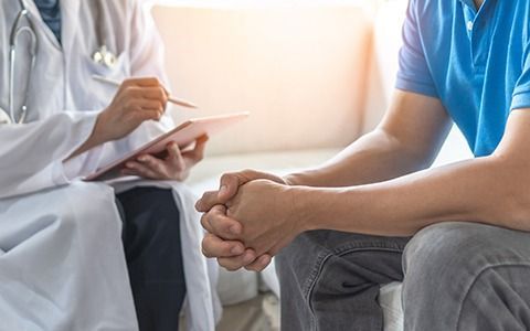 Doctor in white coat taking notes; patient in blue shirt sits with hands clasped, light background.