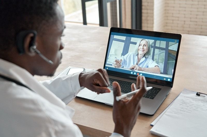 Doctor in white coat on video call with another doctor, gesturing and looking at laptop.