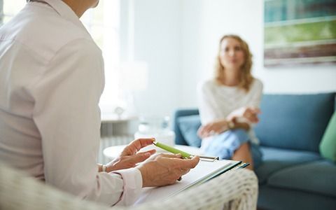 Therapist in white coat taking notes during a session with a woman on a blue couch.
