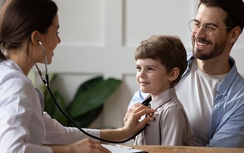 Doctor using a stethoscope on a child patient while the father smiles; in an office setting.