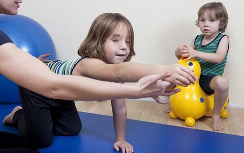 Girl on hands and knees, assisted by an adult, another child on a yellow toy in background, blue mat, indoor.