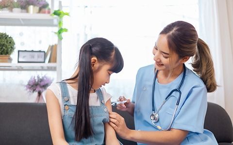 Nurse giving a young girl a shot in her arm, both smiling. Indoors, seated on a couch.