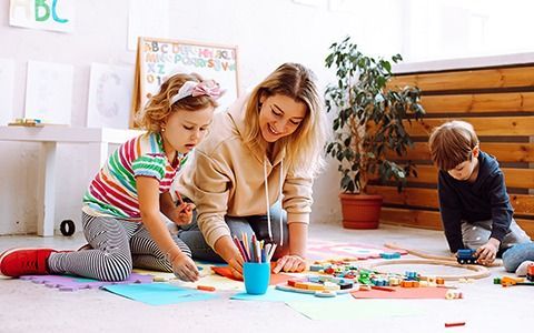 Woman and children playing with art supplies on the floor in a brightly lit room.