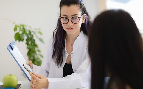 A nutritionist in a white coat reviews a clipboard with a client. A green apple is on the desk.