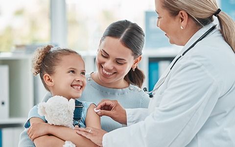 Child receiving a vaccine from a doctor, held by her smiling mother in a clinic.