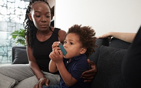 Woman helps child use an inhaler on a couch. Both have concerned expressions.