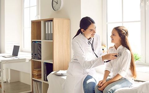 A doctor listens to a young girl's chest with a stethoscope in a sunny office.