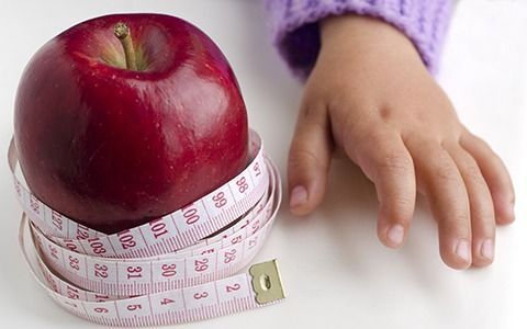 Red apple wrapped with measuring tape, next to a child's hand in a purple sweater.