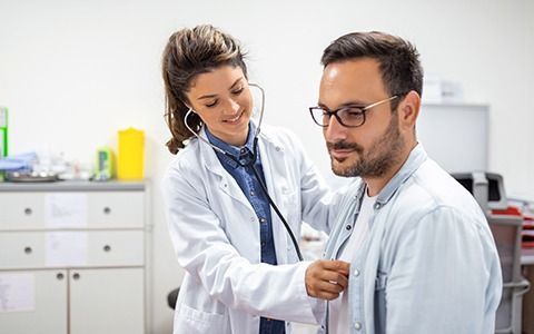 Female doctor listens to male patient's chest with stethoscope in a clinic; smiling.