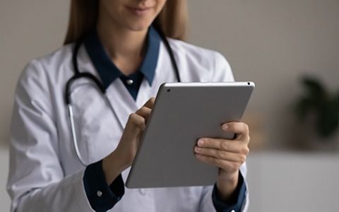 Doctor in white coat and stethoscope uses tablet. Smiling, indoors.