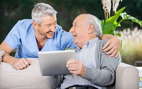 Man with silver hair smiles with an elderly man, holding a tablet, outdoors.