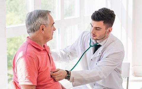 Doctor using stethoscope on older patient's chest in a bright room.