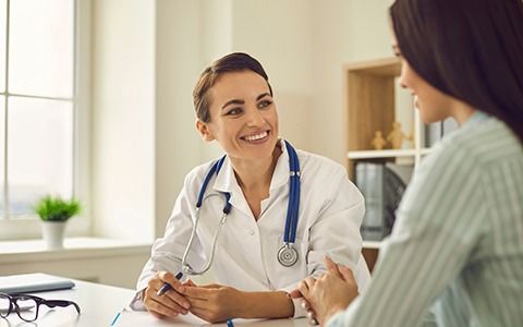 A smiling doctor with stethoscope talks to a patient in a bright office setting.