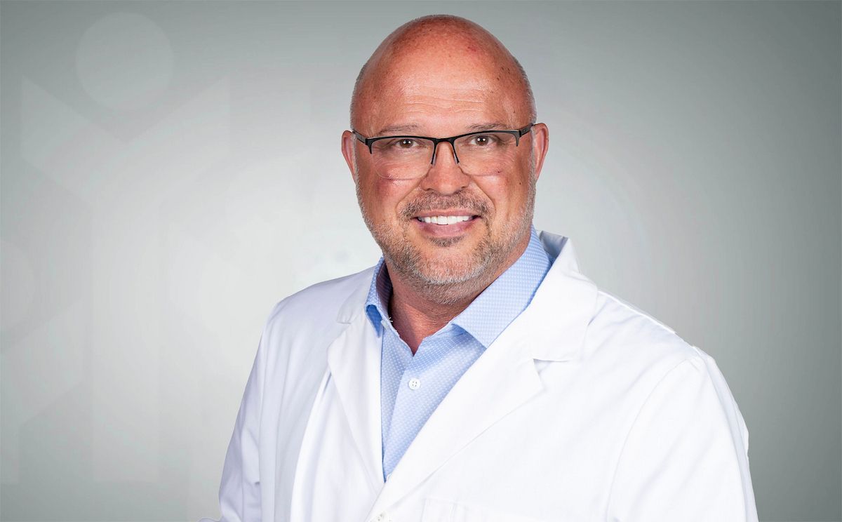 Todd Greene-PA-C.Smiling bald man in glasses wearing a white lab coat against a gray background