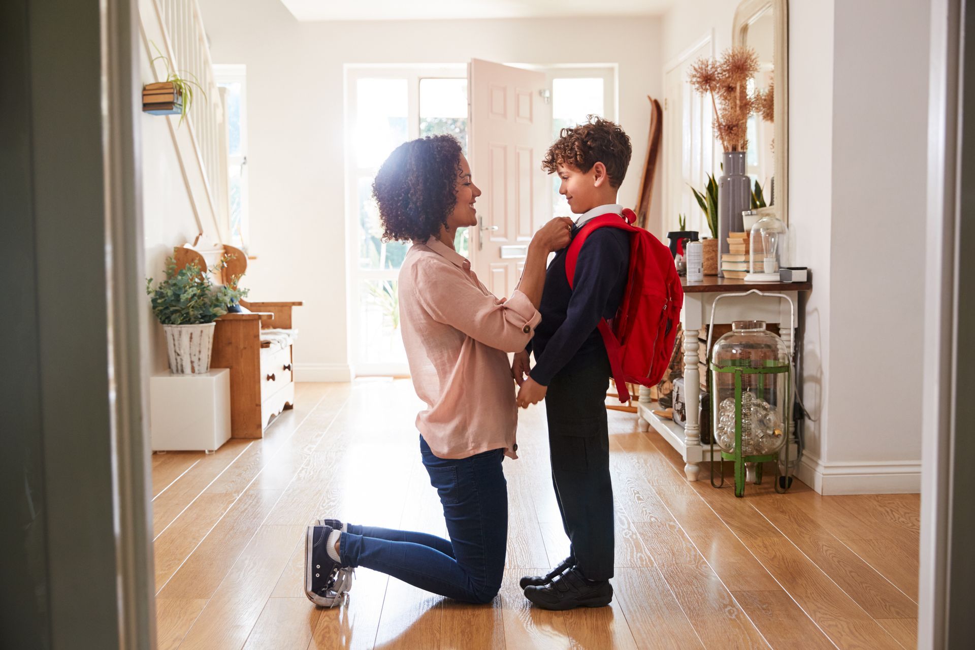 Woman kneels, adjusting a young boy's backpack in a bright hallway. They face each other, ready for school.