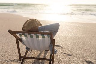 Person relaxing in a beach chair, facing the ocean, wearing a hat. Sandy beach, bright sunlight.