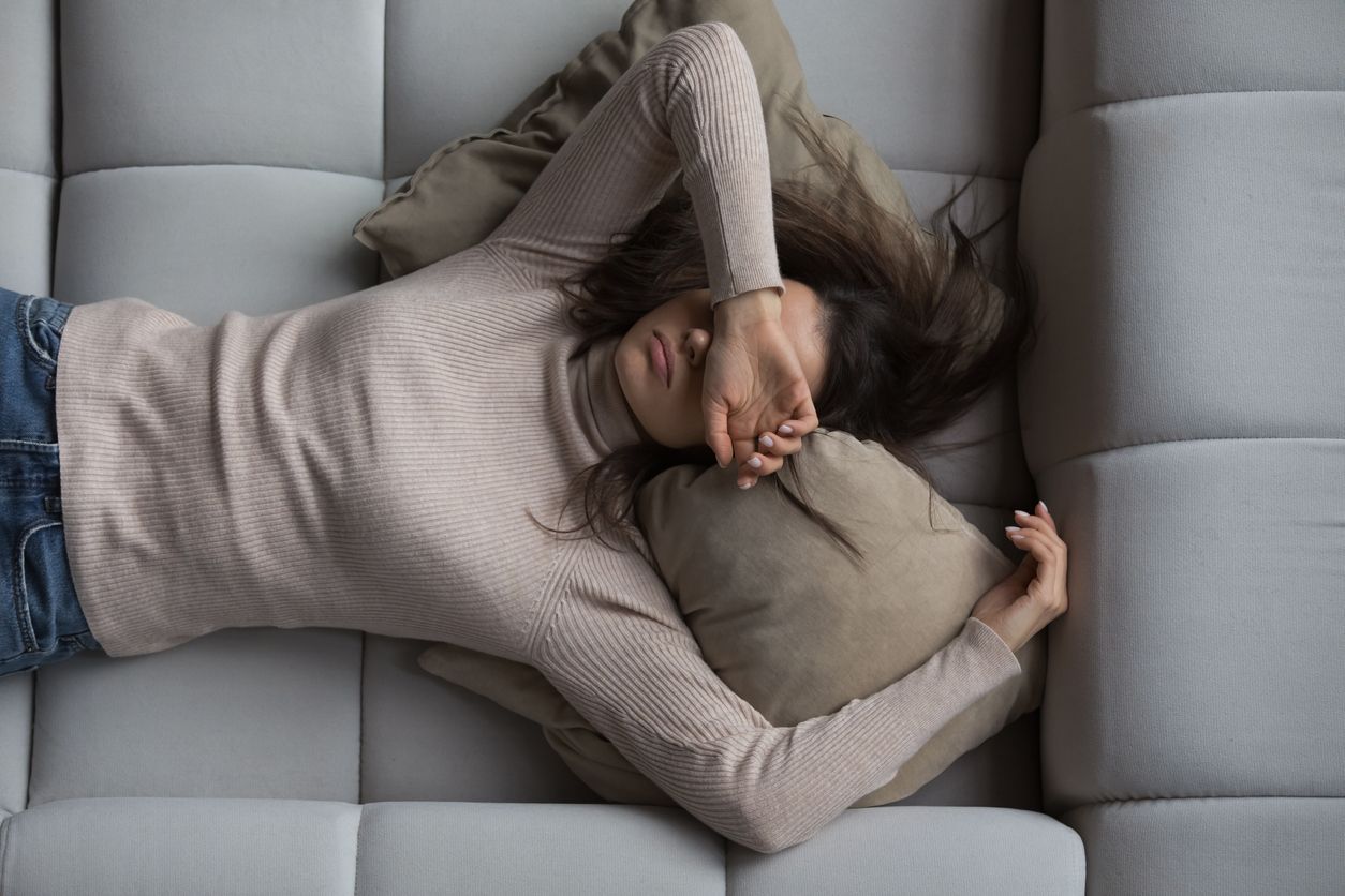 Woman lying on gray couch, covering face with hand, appearing tired.