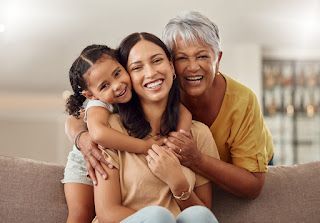 Three generations of smiling women hugging on a couch; a girl, mother, and grandmother.