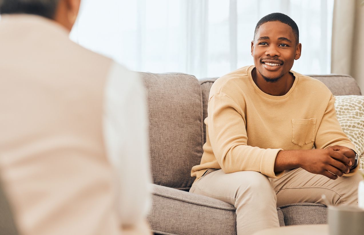 Young Black man smiling on a couch, facing an older person with their back to the viewer in a living room.