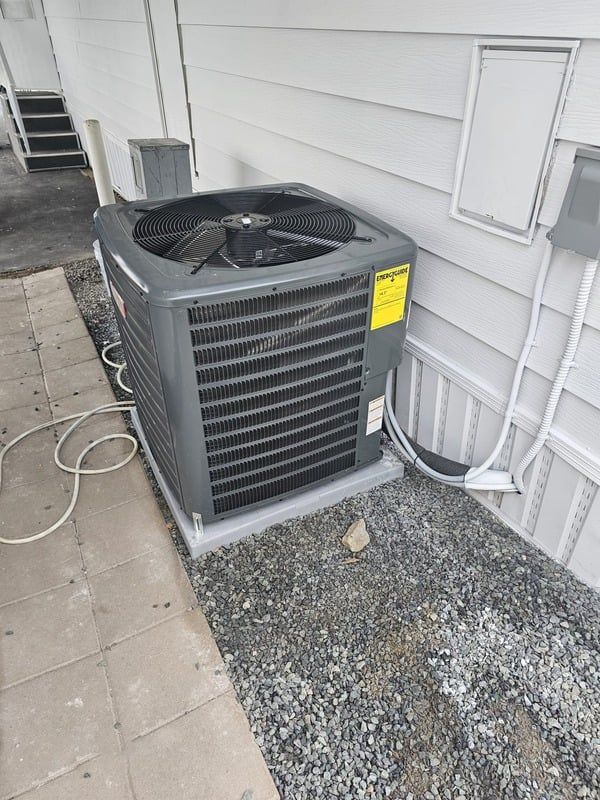 Outdoor air conditioner unit beside a house wall on a gravel pad, with hoses and electrical conduit nearby.