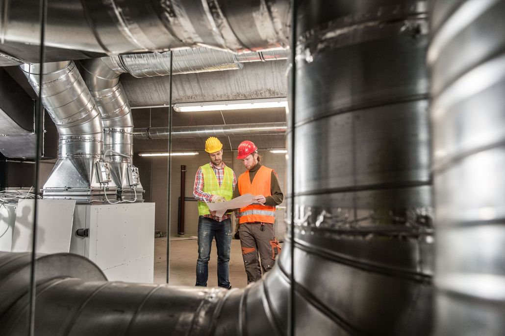 Two workers in hard hats and safety vests inspecting large HVAC ducts in an industrial room