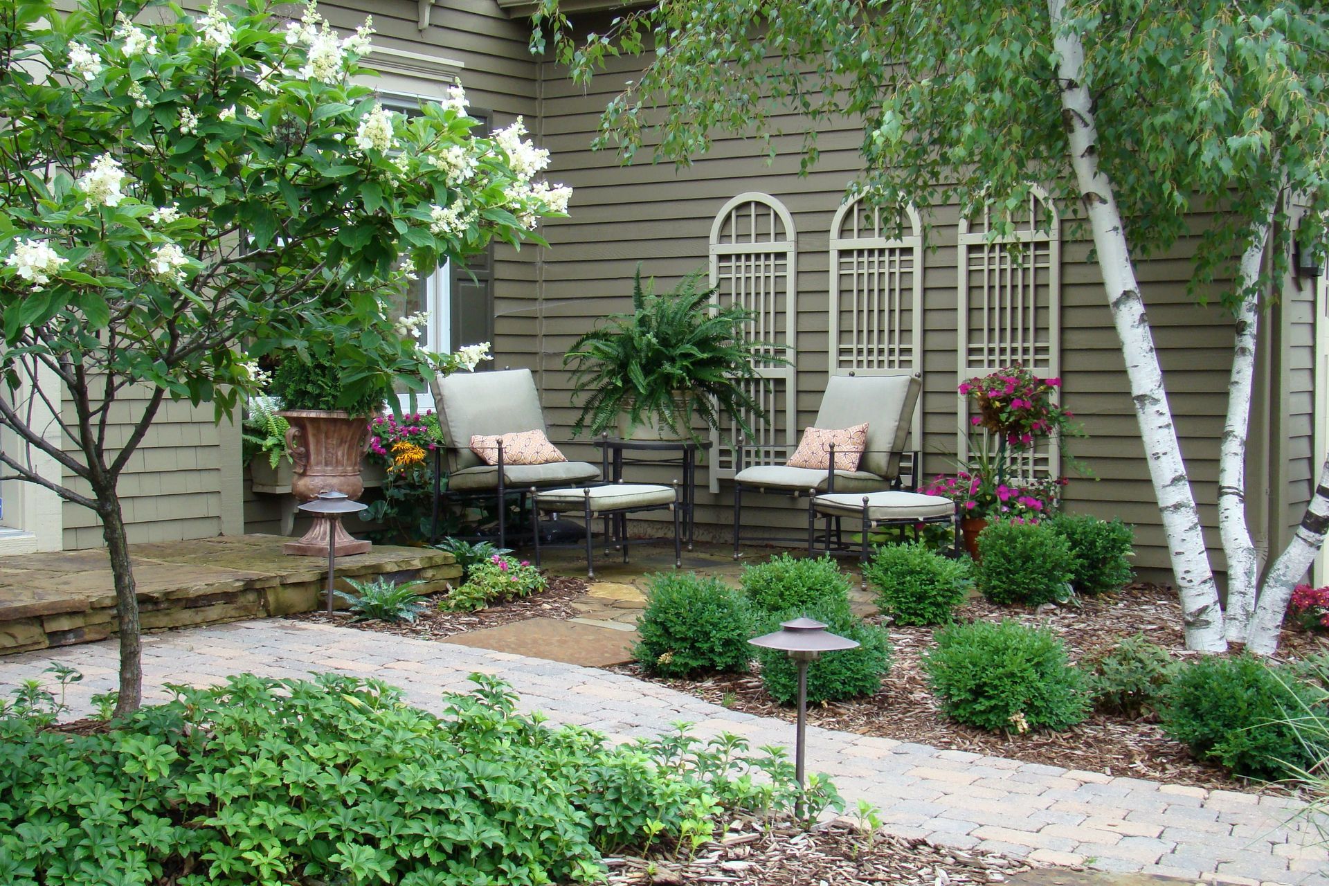 Cozy outdoor seating area with chairs, small table, and decorative trellises. Lush greenery and paved pathway.