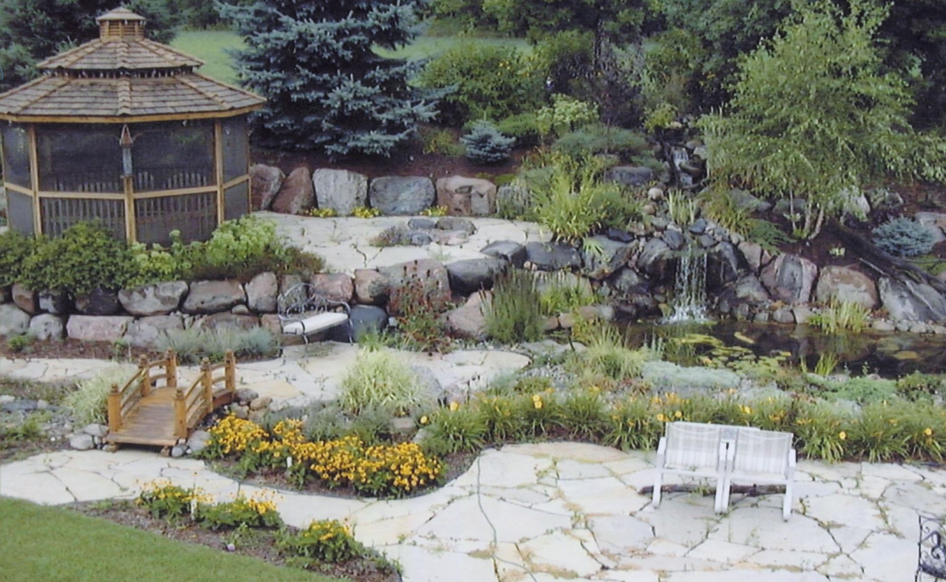 Gazebo, waterfall, and pond in a landscaped garden with stone pathways and a wooden bench.