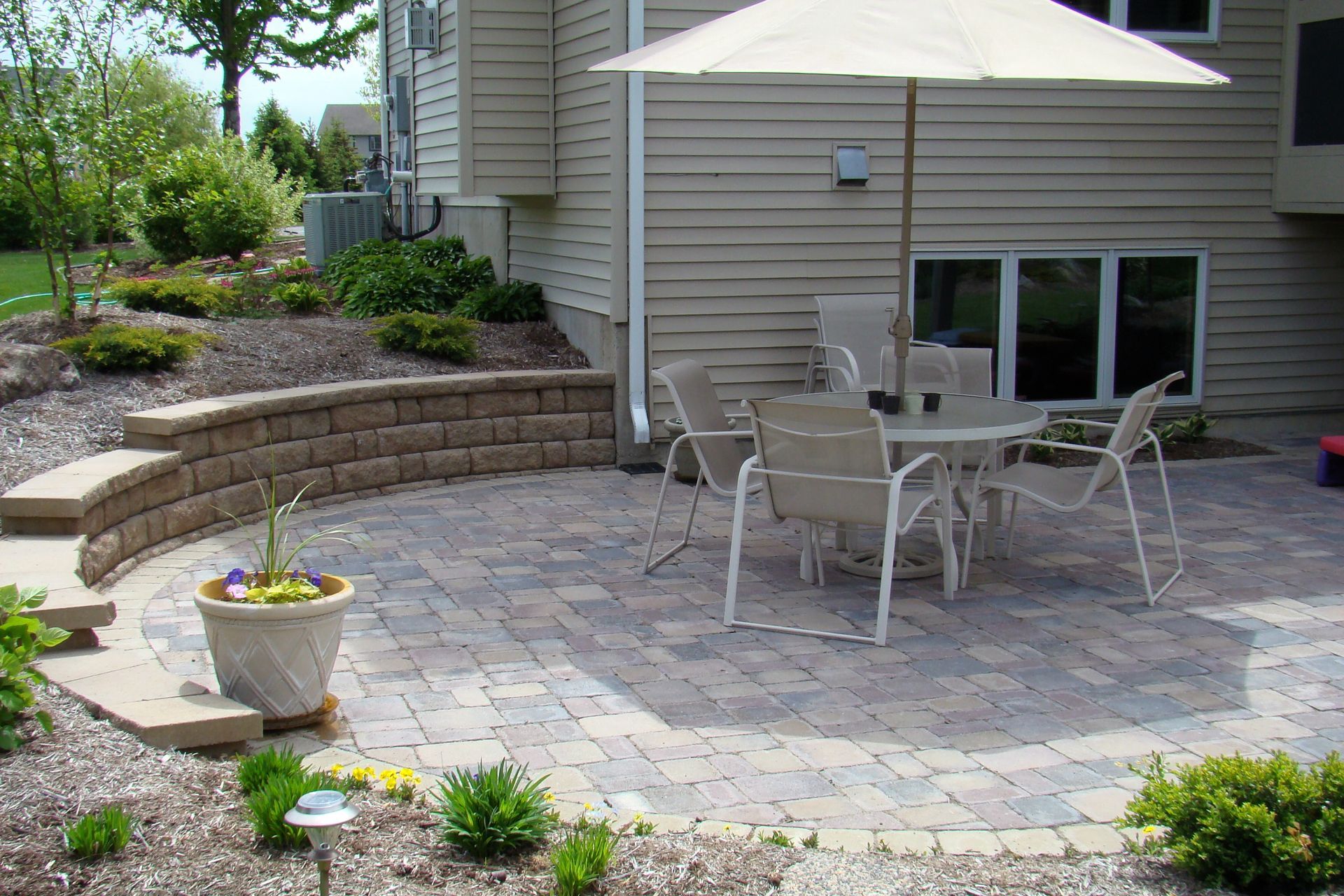 Patio with a table and chairs under an umbrella, surrounded by landscaping and a low retaining wall.