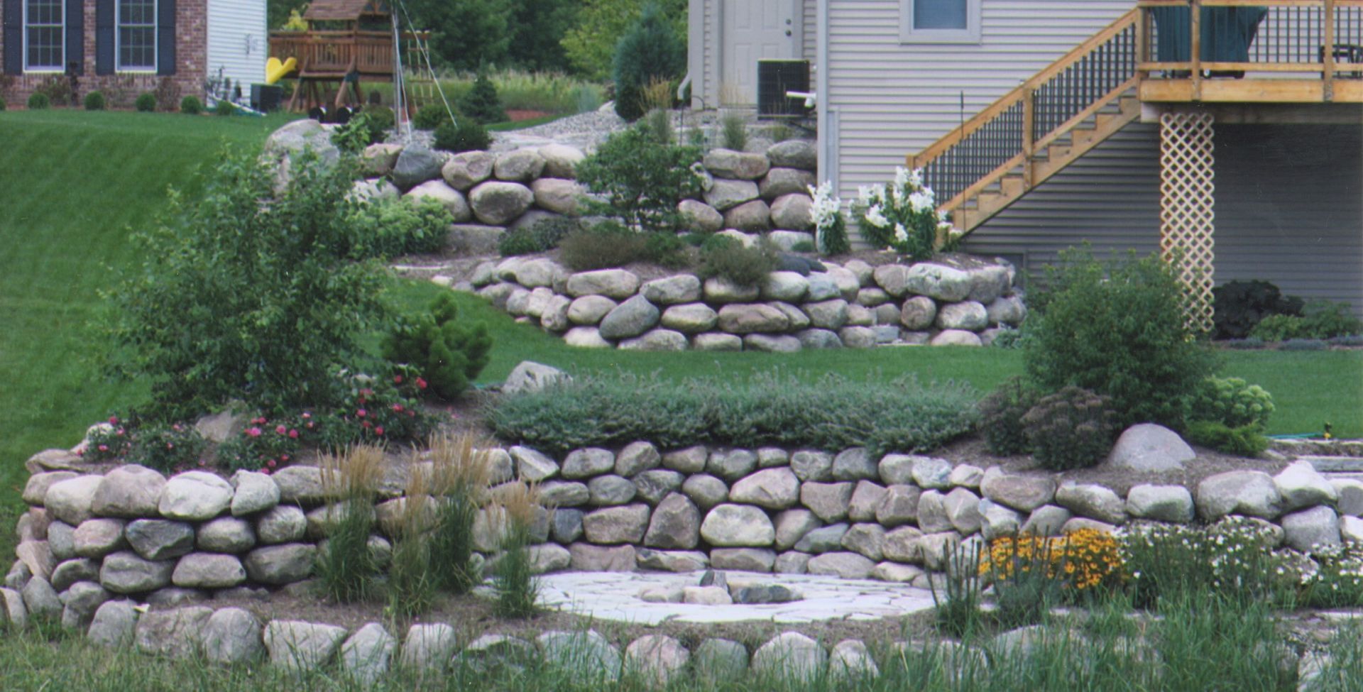 Stone terraced hillside garden with plants and stairs leading to a house deck.