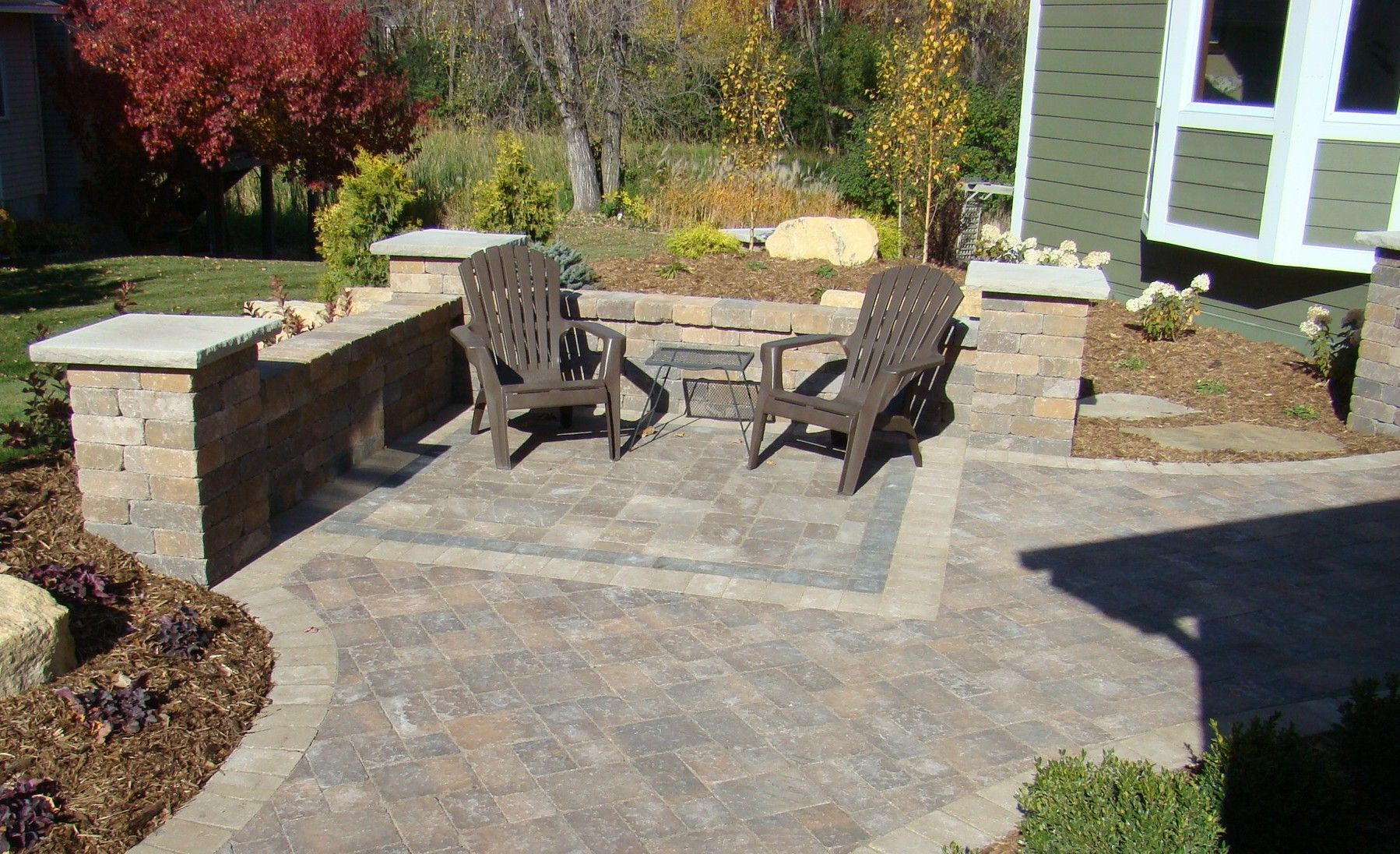 Patio with stone pavers, chairs, and low stone walls. Autumn foliage in the background.
