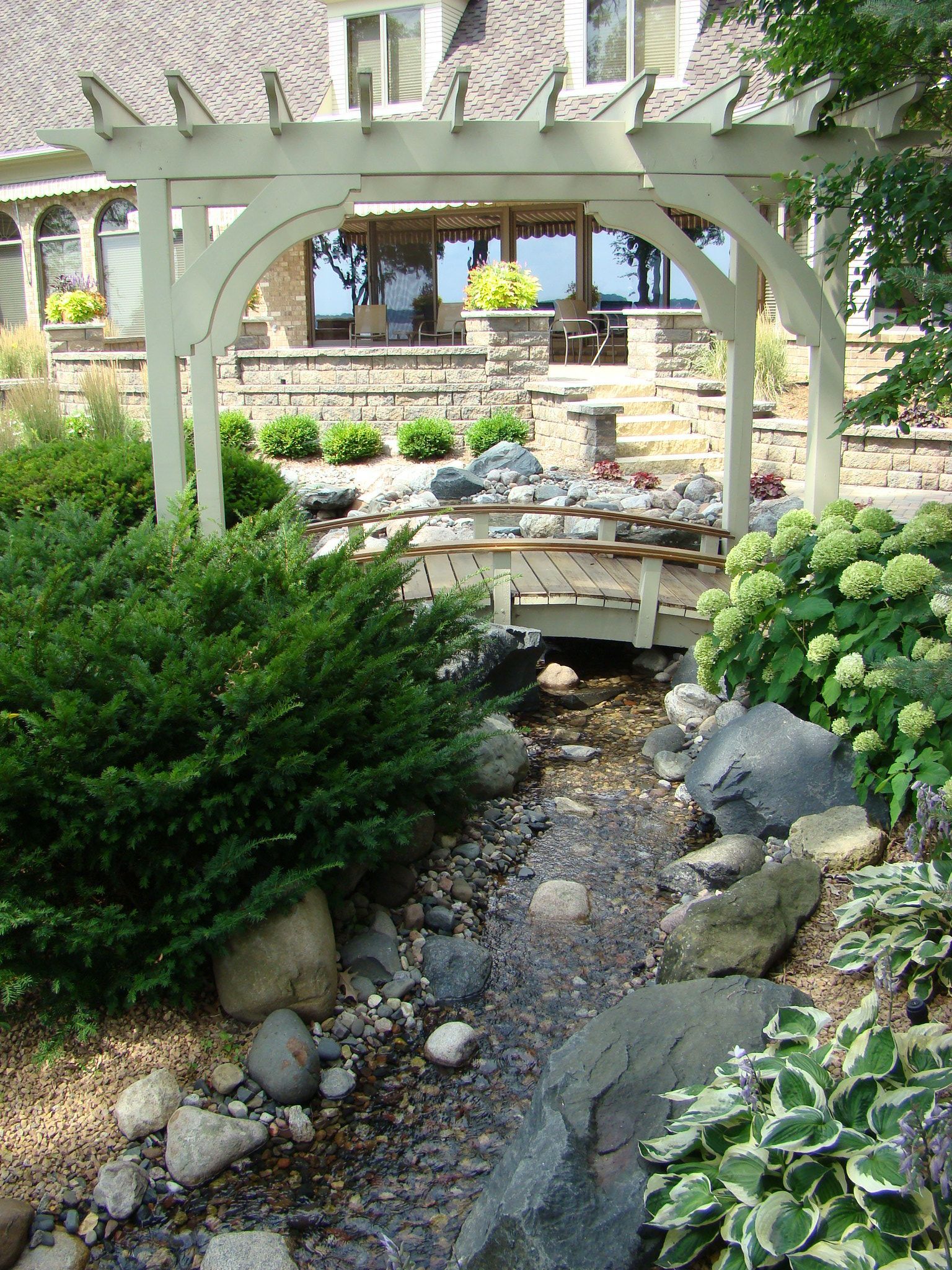 Arched white pergola frames a small stream flowing through a garden with rocks and green plants, near a house.