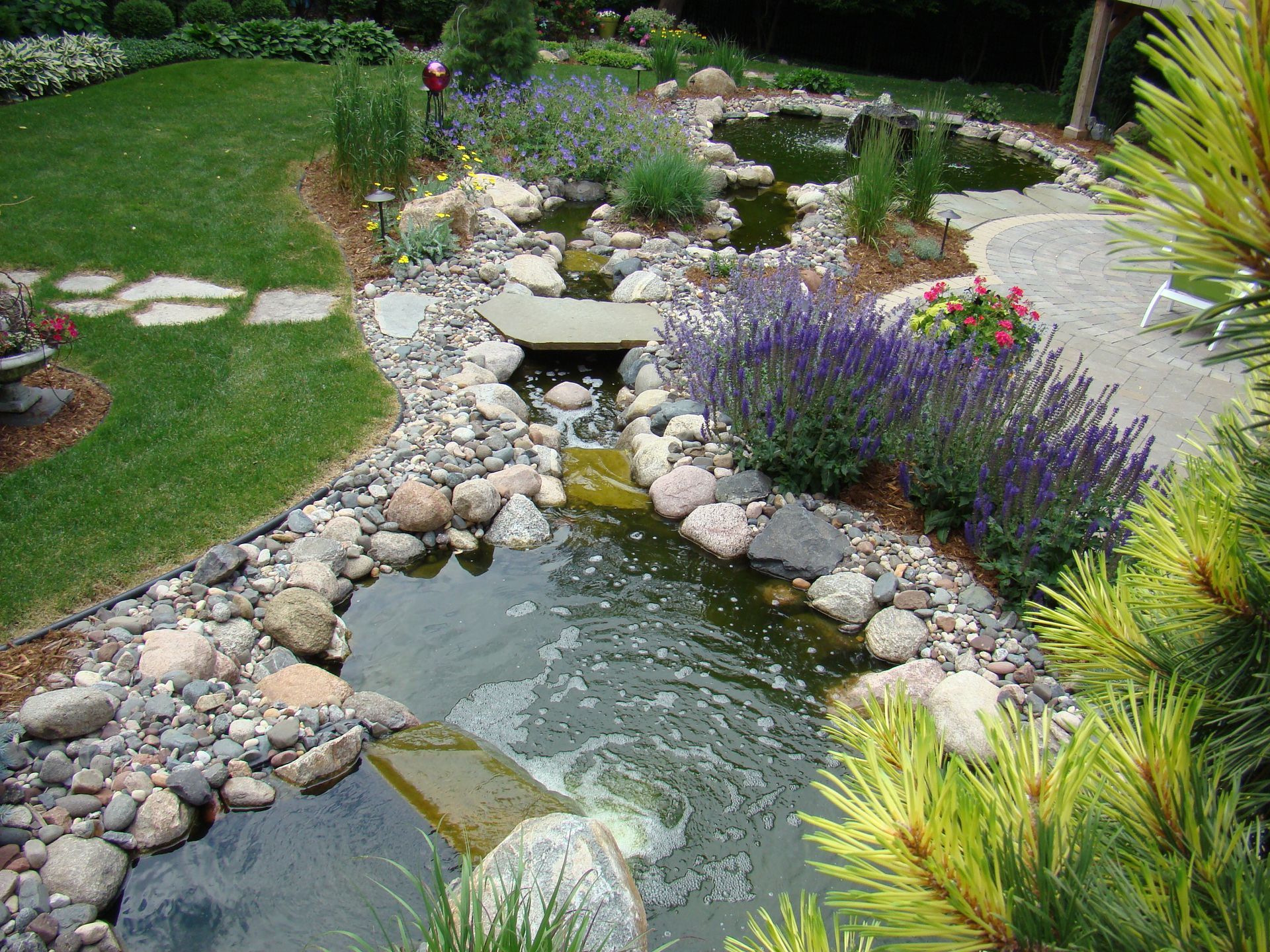 Stone water garden with cascading water, rocks, and purple flowers. Lush green grass and shrubs surround it.