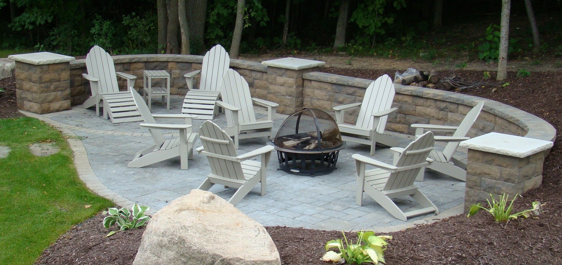A backyard fire pit surrounded by Adirondack chairs, stone retaining walls, and landscaping.