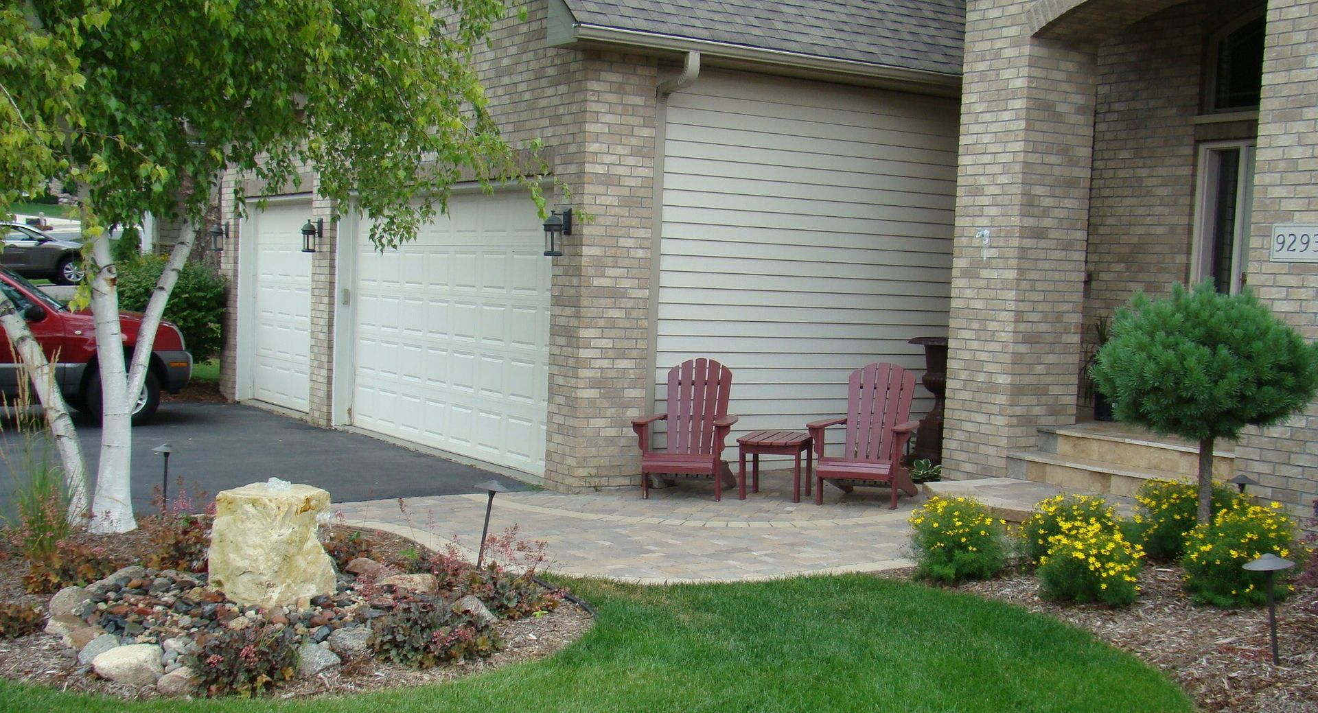 House exterior with beige stone facade and three white garage doors. Red chairs and a small table sit on a patio.