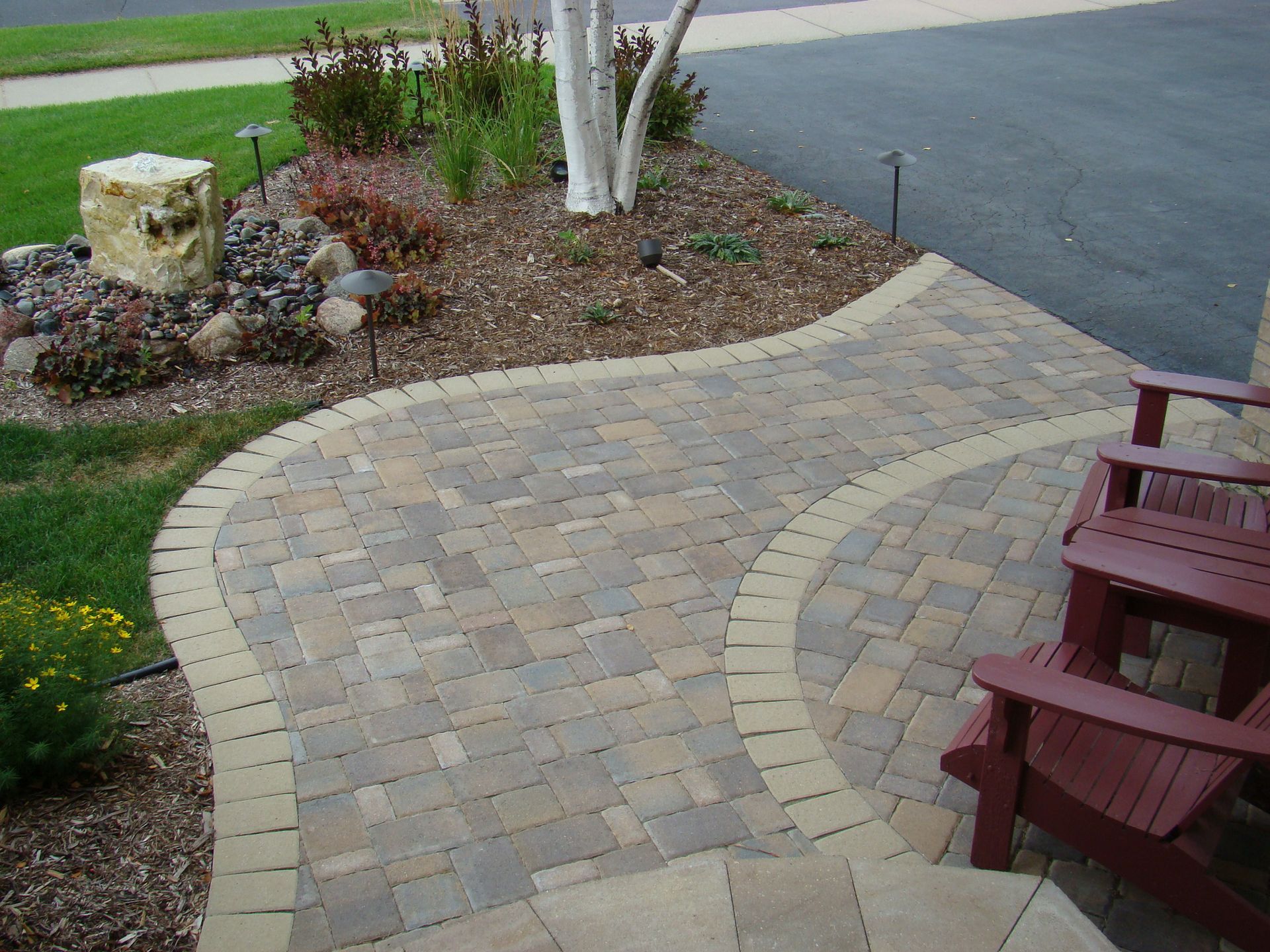 Brick patio with curved border, leading to chairs. Landscaping includes trees, rocks, and mulch.