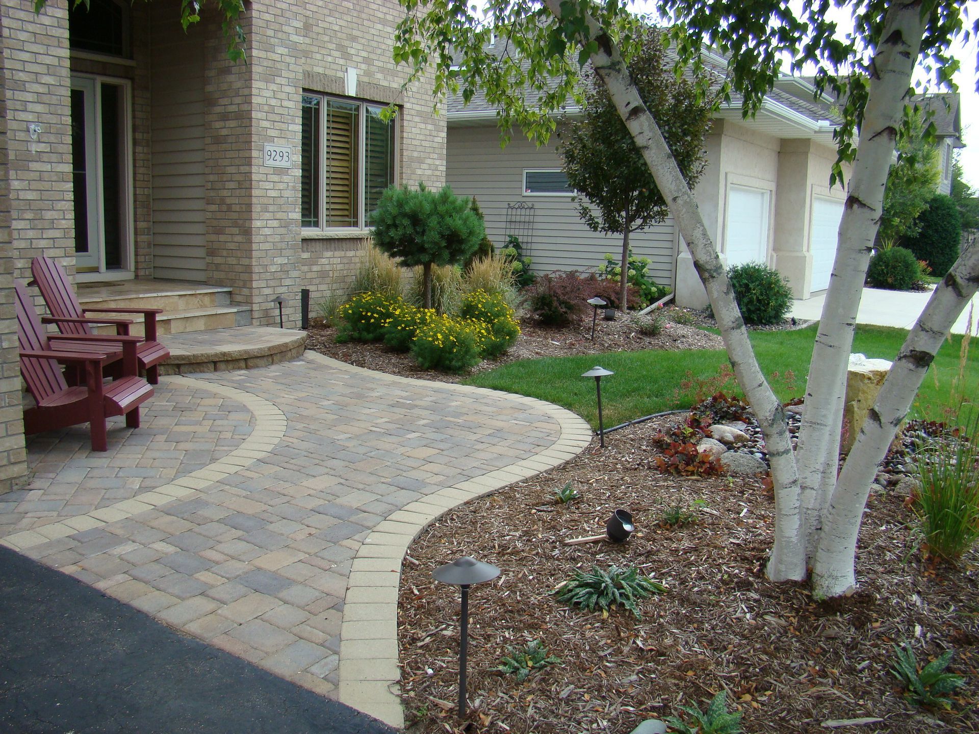 Brick pathway curves towards a house with landscaping, trees, and seating.