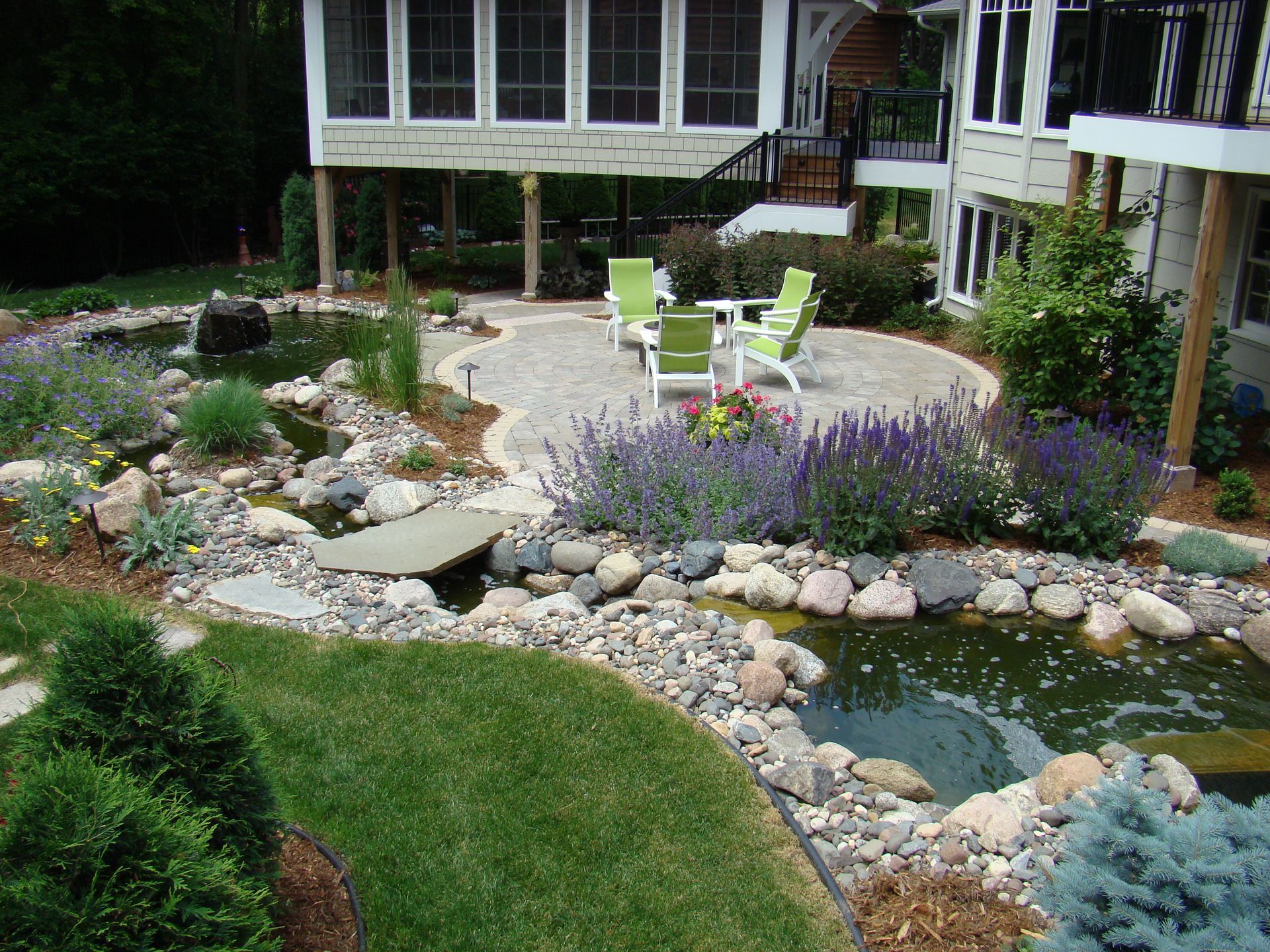 Lush backyard with a stone patio, pond, bridge, and landscaping surrounding a house.